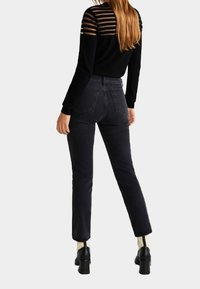 Haut noir à manches longues avec partie supérieure transparente rayée et jean noir ajusté. Le mannequin porte des bottines à talons carrés. Textures de tissu simples et lisses.