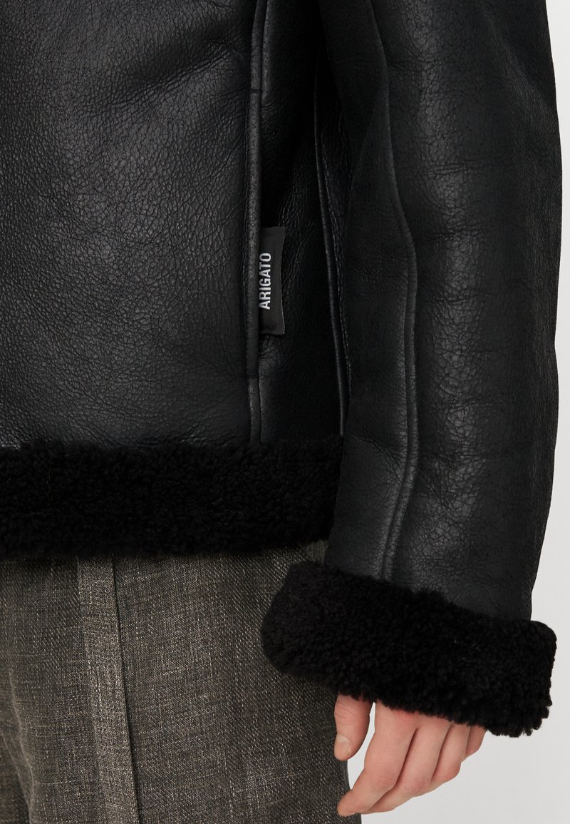 Chaqueta de cuero negra con cuello y puños de borreguillo. Presenta un acabado texturizado, costura lateral y una etiqueta con la inscripción "ARIGATO". Pantalones grises visibles por debajo.