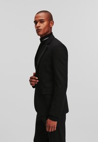 Must värviline ülikond slim-fit lõikega, millel on satiinist krae ja kõrge kaelusega särk. Sile tekstuur, minimalistlik disain ja ilma nähtavate mustriteta.