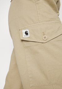 Gros plan sur la poche d’un pantalon Carhartt marron clair avec un rabat boutonné et un écusson du logo Carhartt visible sur la poche.