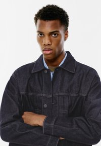 Tamno plavi traper jakna s ovratnikom, prednjim gumbima i vidljivim šavovima. Model nosi plavu košulju ispod; ruke prekrižene ispred.