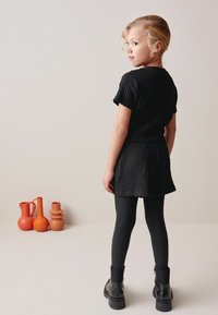 Sort tekstureret kjole med korte ærmer, parret med sorte leggings og kraftige ankelstøvler. Synlige orange keramikvaser i baggrunden.