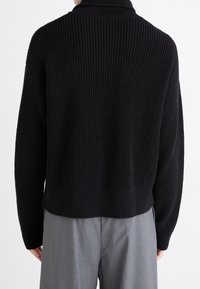 Schwarzer Strickpullover mit gerippter Textur, hoher Kragen und kurzer Saum, kombiniert mit grauen Hosen für eine lässige Passform.