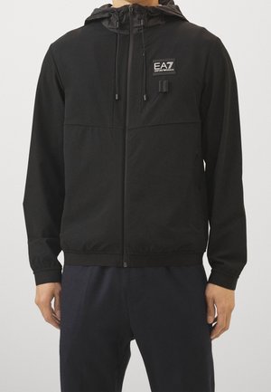 Homme portant une veste à capuche zippée noire avec le logo EA7 Emporio Armani sur la poitrine, associée à un pantalon foncé sur un fond uni.