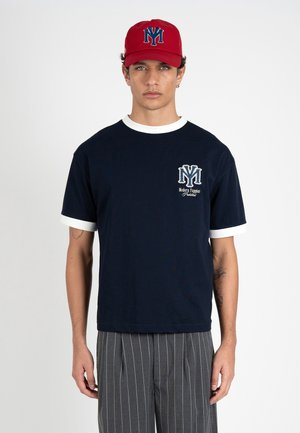 Junger Mann mit roter Kappe und dunkelblauem T-Shirt mit weißem Rand und Logo, kombiniert mit grauen Nadelstreifenhosen, vor weißem Hintergrund stehend.