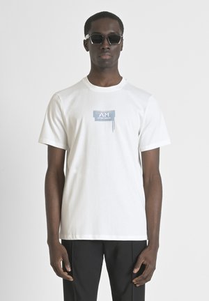 Camiseta de algodón blanca de manga corta, con un estampado gráfico azul cerca del pecho que dice "Anthony Morato", combinada con unos pantalones negros.