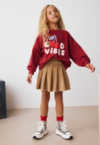 Sweat-shirt rouge avec le texte « GOOD VIBES » et un motif cerise, associé à une jupe plissée beige et des baskets imprimé léopard avec des touches de rouge.