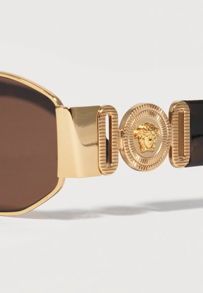 Καφέ γυαλιά ηλίου με χρυσό πλαίσιο και λεπτομερή χρυσό έμβλημα Μέδουσας στον βραχίονα που συνδέεται με μαύρους βραχίονες.