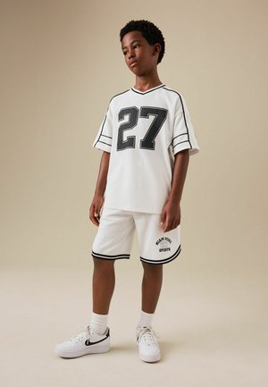 Chlapec stojí a má na sebe biely športový dres s číslom 27 a zodpovedajúce šortky s čiernym lemovaním a biele tenisky, béžové pozadie.