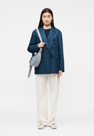 Γυναίκα που φοράει μπλε σακάκι διπλής σειράς κουμπιών, άσπρο παντελόνι με φαρδύ πόδι, μπεζ παπούτσια και κρατάει μία ανοιχτό γκρι τσάντα ώμου.