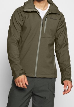 Homme portant une veste zippée Columbia vert olive avec poches sur la poitrine et les côtés, associée à un pantalon gris foncé.