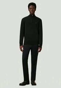 Homem de pé, vestindo um suéter preto com fecho, calças pretas e sapatos pretos, contra um fundo claro e simples.
