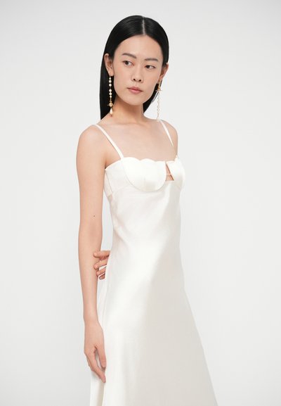 Žena s dlhými čiernymi vlasmi, oblečená v bielych saténových šatách s tenkými ramienkami a výraznými dlhými perlovými náušnicami, pózujúca pred jednoduchým pozadím.