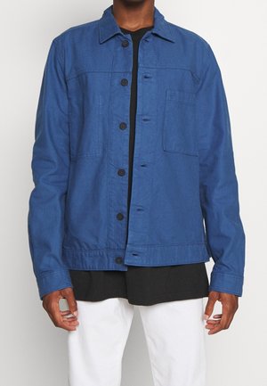 Mann trägt eine blaue, knöpfbare Jacke über einem schwarzen Hemd mit weißen Hosen, steht vor einem schlichten hellen Hintergrund.