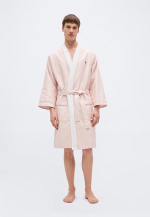 Jonge man staat zonder schoenen, gekleed in een lichtroze met witte gestreepte, knielange badjas met een ceintuur om de taille geknoopt.