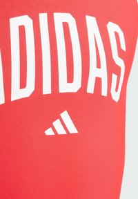 Červené sportovní oblečení s velkým bílým logem "ADIDAS" a malým designem tří pruhů. Hladká, pružná tkanina s jednobarevným pozadím.