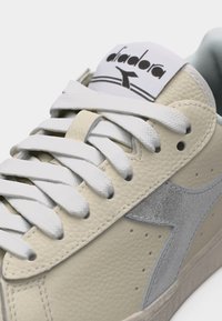 Κοντινό πλάνο μπεζ αθλητικού παπουτσιού Diadora με λευκά κορδόνια και γκρι λογότυπο από σουέτ σε κεντρικό δερμάτινο πάνω μέρος με υφή.