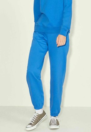 Joggers de algodón azules con puños elásticos, bolsillos laterales y un corte relajado, combinados con zapatillas grises. Textura suave y color brillante.