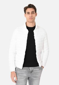 Giacca bianca trapuntata con colletto alto e chiusura con zip, abbinata a un maglione nero e jeans grigi, su uno sfondo chiaro.