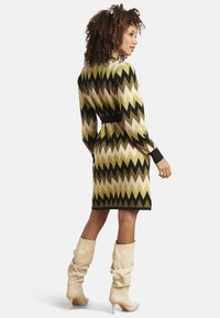 Vestito a maglia con motivo a zigzag in tonalità di verde, beige e nero. Vita aderente, maniche lunghe e polsini a costine. Lunghezza al ginocchio.