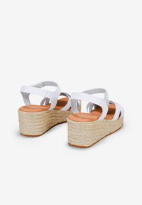Sandalias de cuña blancas con múltiples correas entrelazadas, plataforma de yute texturizado y una plantilla de cuero suave. Presenta una suela de color tostado y bordes redondeados.