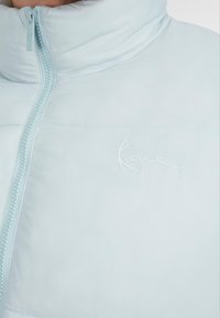 Šviesiai mėlynas pūkinis švarkas su aukštu apykakle, užtrauktuku ir subtiliu išsiuvinėtu logotipu. Lygus paviršius su grakščiu dizainu.