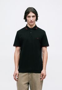 Young man with medium-length hair wearing a black polo shirt with red trim and beige pants, standing against a plain white background.