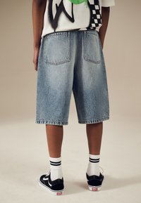 Jeans-Shorts in Hellblau, knielang mit lockerem Schnitt, versehen mit zwei Gesäßtaschen und einem ausgefransten Saum, kombiniert mit schwarzen Turnschuhen und weißen gestreiften Socken.