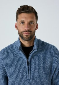 Gaiši zila adīta džemis ar augstu apkakli un pusvilkšanu, ar strukturētu rakstu un ribotām akcentiem.