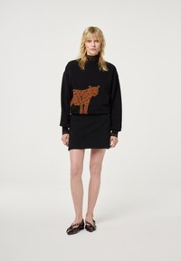 Blonde Frau steht mit einem schwarzen Pullover mit großem braunem Leopardenmuster, einem schwarzen Cordminirock und dunkelroten spitzen Ballerinas mit Riemchen.