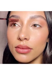 A person applying brown mascara with a wand on their eyelashes. They have smooth skin and glossy light pink lips.