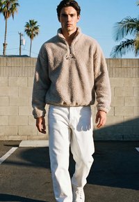 Fiatal férfi sétál kint, bézs polár pulóvert, fehér nadrágot és fehér tornacipőt viselve, pálmafák és téglafal háttér előtt.