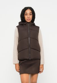 Giubbotto imbottito marrone con cappuccio, dotato di chiusura frontale con zip e cuciture diagonali. Indossato sopra una maglia a maniche lunghe beige chiaro.
