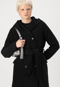 Abrigo de lana negro con capucha, diseño de doble botonadura, botones dorados y cinturón. Bolso plateado con acabado pulido y correa ajustable.