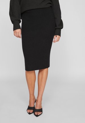 Kvinde iført sort ribstrikket pencilnederdel, sort langærmet sweater og sorte åbne hæle, stående på en grå gulv.