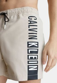 Costumi da bagno beige con logo "CALVIN KLEIN" nero stampato verticalmente lungo il lato, vita elasticizzata e chiusura con coulisse.