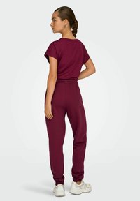 Bordo dvodijelni outfit s kratkim rukavima i elastičnim pojasom. Sužene hlače za trčanje s manžetama uz članak. Model nosi bijele tenisice.
