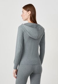 Mujer vistiendo una sudadera gris ajustada con capucha y leggings gris a juego, de espaldas contra un fondo blanco liso.
