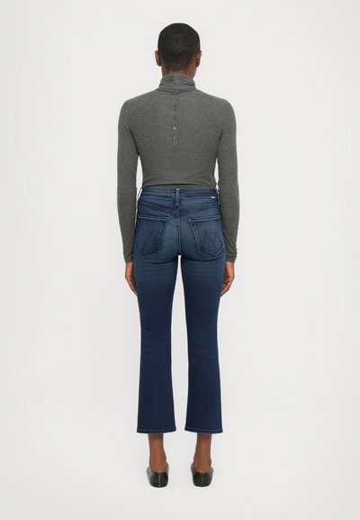 Pulover gri cu guler înalt, cu detalii cu butoane, și blugi evazați de culoare albastră închis, cu buzunare pe spate, purtați cu pantofi negri. Design simplu, ajustat.