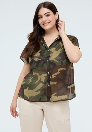Femme souriante portant une chemise boutonnée à manches courtes camouflage vert et marron et un pantalon crème, ajustant une grande boucle d'oreille avec la main près du visage.