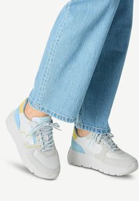 Sneakers bianchi e azzurri chiari con accenti gialli, caratterizzati da una suola spessa, chiusura con lacci e materiali superiori testurizzati. Abbinati a jeans azzurri chiari.