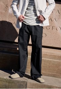 Osoba w czarnych spodniach, beżowych butach na slip-on, w pasiastym szaliku i białej marynarce stoi przy kamiennej ścianie z wyrytym tekstem.