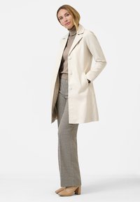 Crème-kleurige jas met een op maat gemaakt ontwerp, knoopsluiting en zakken, gecombineerd met een bruine coltrui en broek met houthakkerspatroon.