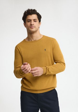 Man in mosterdgele trui met klein donker logo die zijn mouw aanpast, staand tegen een effen lichte achtergrond.