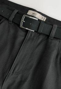 Juodos spalvos raumenų stiliaus siauri kelnės iš tekstūruotos medžiagos, su juoda austa diržu ir sidabro spalvos metaline sagtimi, parodytos kampu.