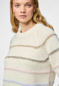 Mlada žena s plavom kosom koja nosi bijeli pleteni pulover s horizontalnim pastelnim prugama i malim srebrnim округлим naušnicama.