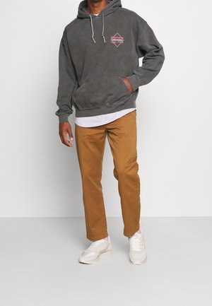 Persona con una sudadera gris oscuro con texto rosa, camiseta blanca, pantalones beige y zapatillas blancas, de pie con una mano en el bolsillo de la sudadera.