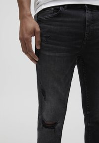 Närbild av en person som bär svarta slitna jeans med små revor på låret och knäet, handen avslappnad vid sidan.