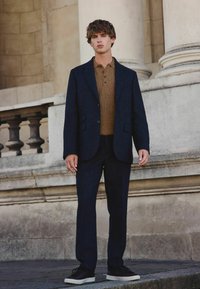 Dunkelblaues Blazer mit Reverskragen über einem braunen Strickpolo, kombiniert mit passenden Hosen und schwarzen Turnschuhen; glatte Textur und taillierte Passform.