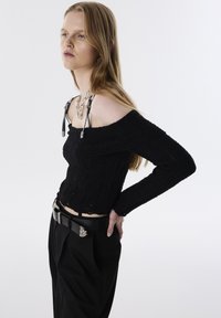 Zwarte gebreide off-shoulder top met lange mouwen en een gestructureerde stof, gecombineerd met hoge zwarte broeken en een riem met zilveren details.